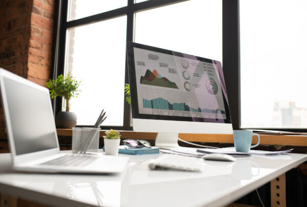 Computer monitor with graphical information on its screen, laptop, mug and other supplies on desk by office window